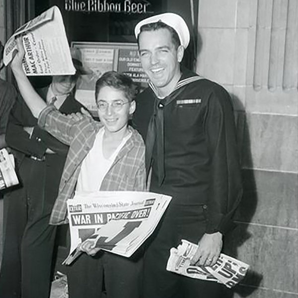 Seaman 1st class Tom Teeley with newsboy, Bernard Ehrmann, holding a copy of the Wisconsin State Journal with headline declaring peace in the Pacific. Celebrating V-J Day, August 15, 1945, the day on which the Allies announced the surrender of Japanese forces during World War II.